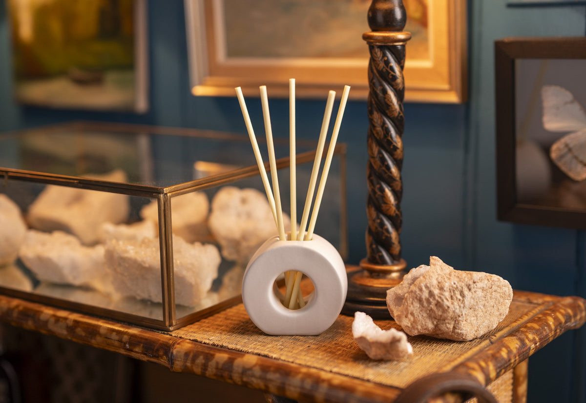 A white ceramic reed diffuser with light wood reeds sits on a decorative wooden table, styled with natural stones and framed wall art in a blue room setting.