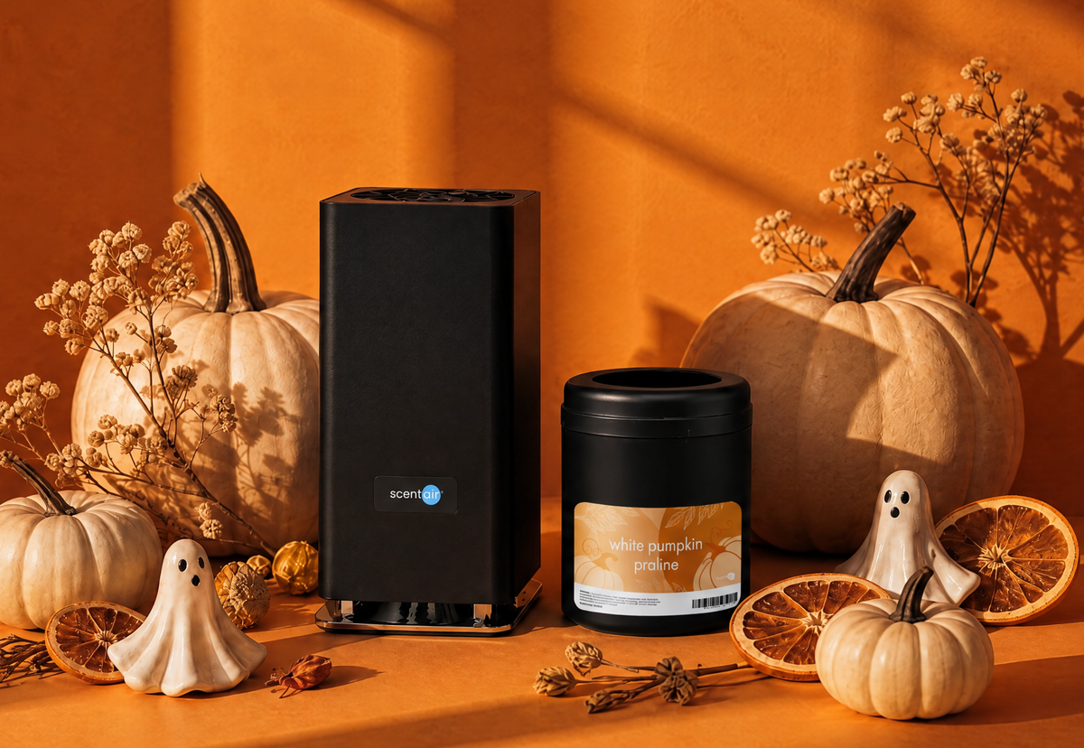 A black ScentAir Whisper PRO fragrance diffuser sets next to a White Pumpkin Praline Whisper PRO fragrance cartridges. They are surrounded by pumpkins, porcelain ghosts, orange slices, and other fall themed decorations with an orange background.