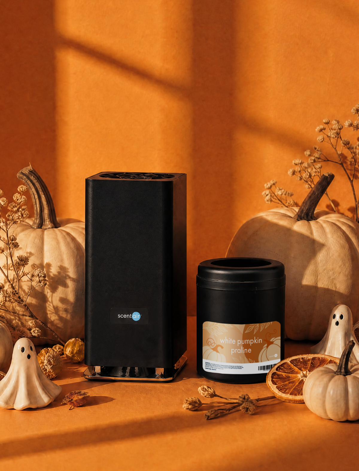 A black ScentAir Whisper PRO fragrance diffuser sets next to a White Pumpkin Praline Whisper PRO fragrance cartridges. They are surrounded by pumpkins, porcelain ghosts, orange slices, and other fall themed decorations with an orange background.