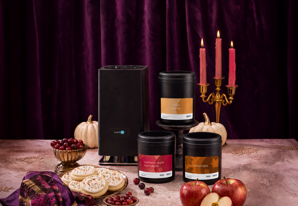 Three black Whisper PRO Cartridges with colorful labels are arranged on a tabletop next to a Whisper PRO diffuser in a styled scene with pumpkins, apples, cranberries, a candle holder, and a dark purple velvet curtain backdrop.