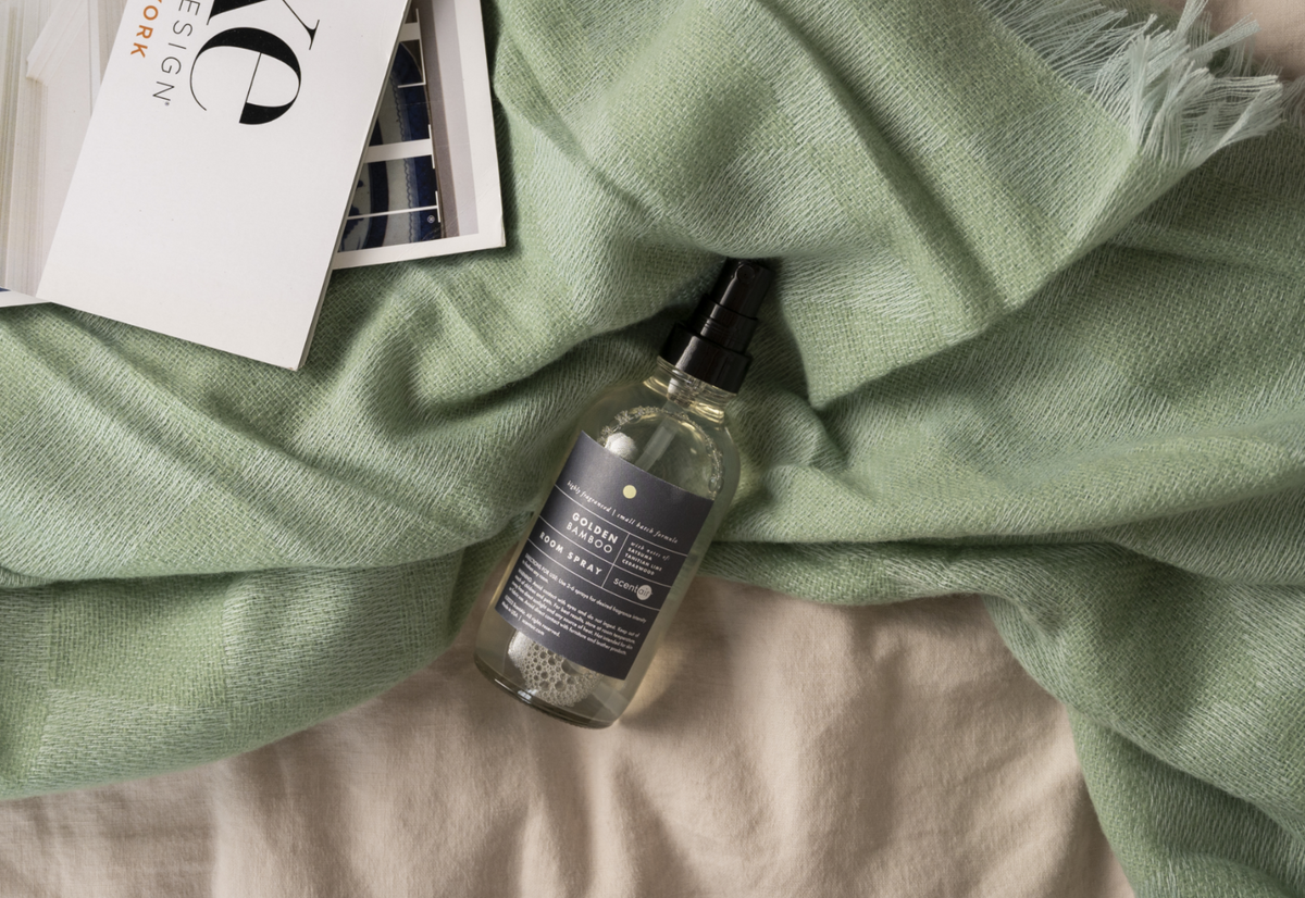 A clear spray bottle with a black pump and dark label lies on a soft sage green throw blanket over a light beige fabric surface. A white card with large black lettering is partly visible at the top left.